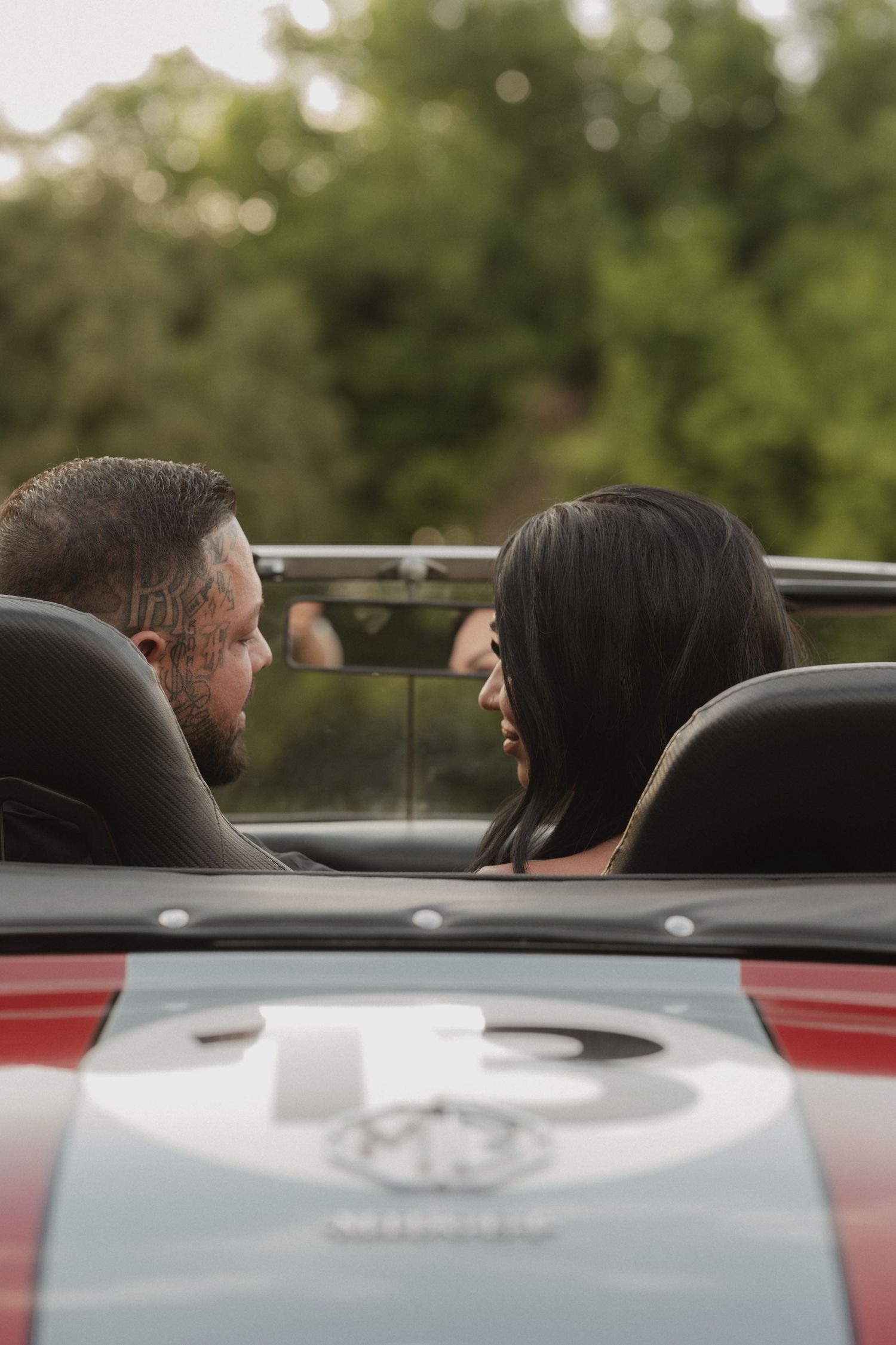A couple sits in a red convertible sports car, gazing at each other romantically, surrounded by lush green trees.