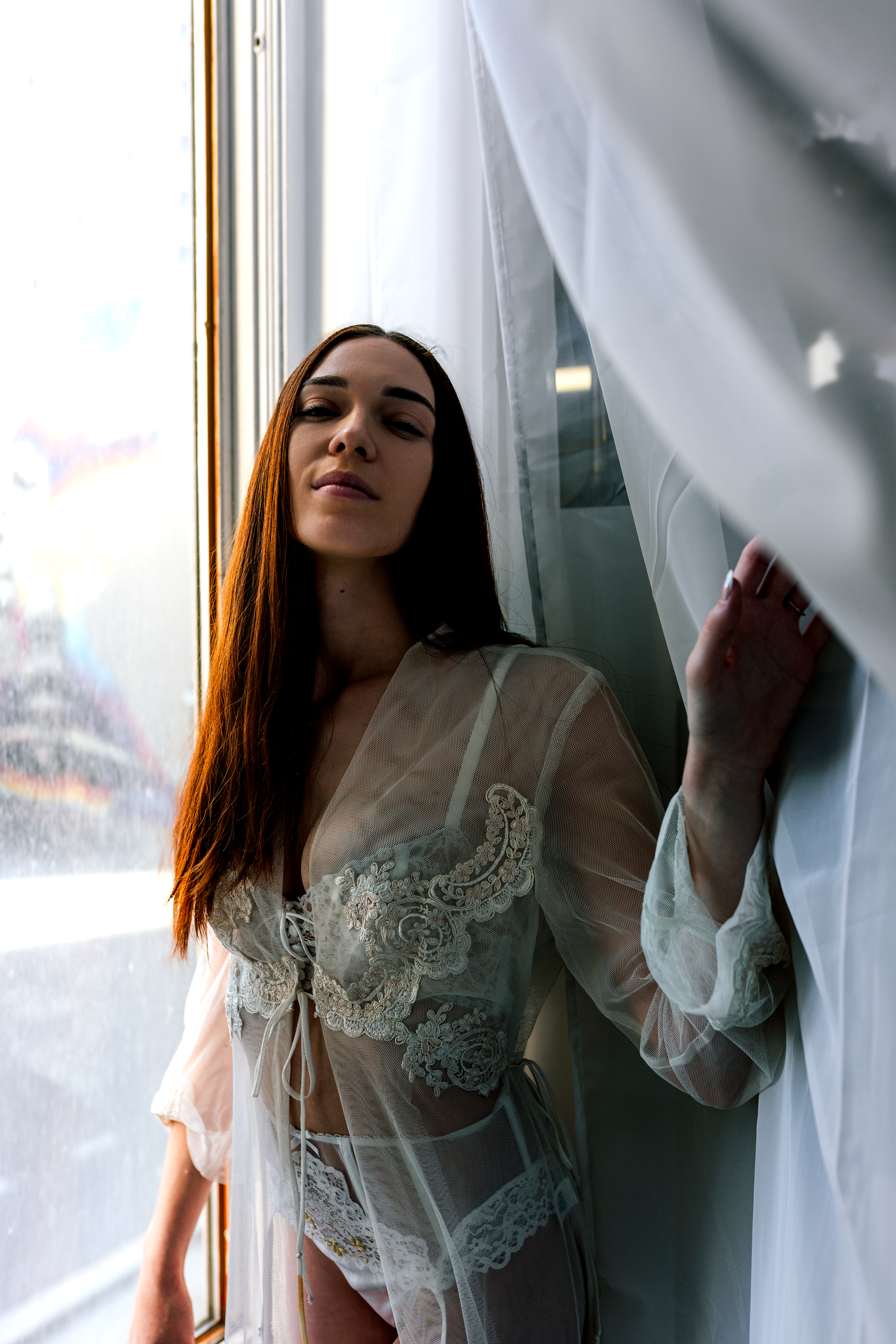Woman in white lace lingerie and sheer robe standing by a window with flowing curtains.