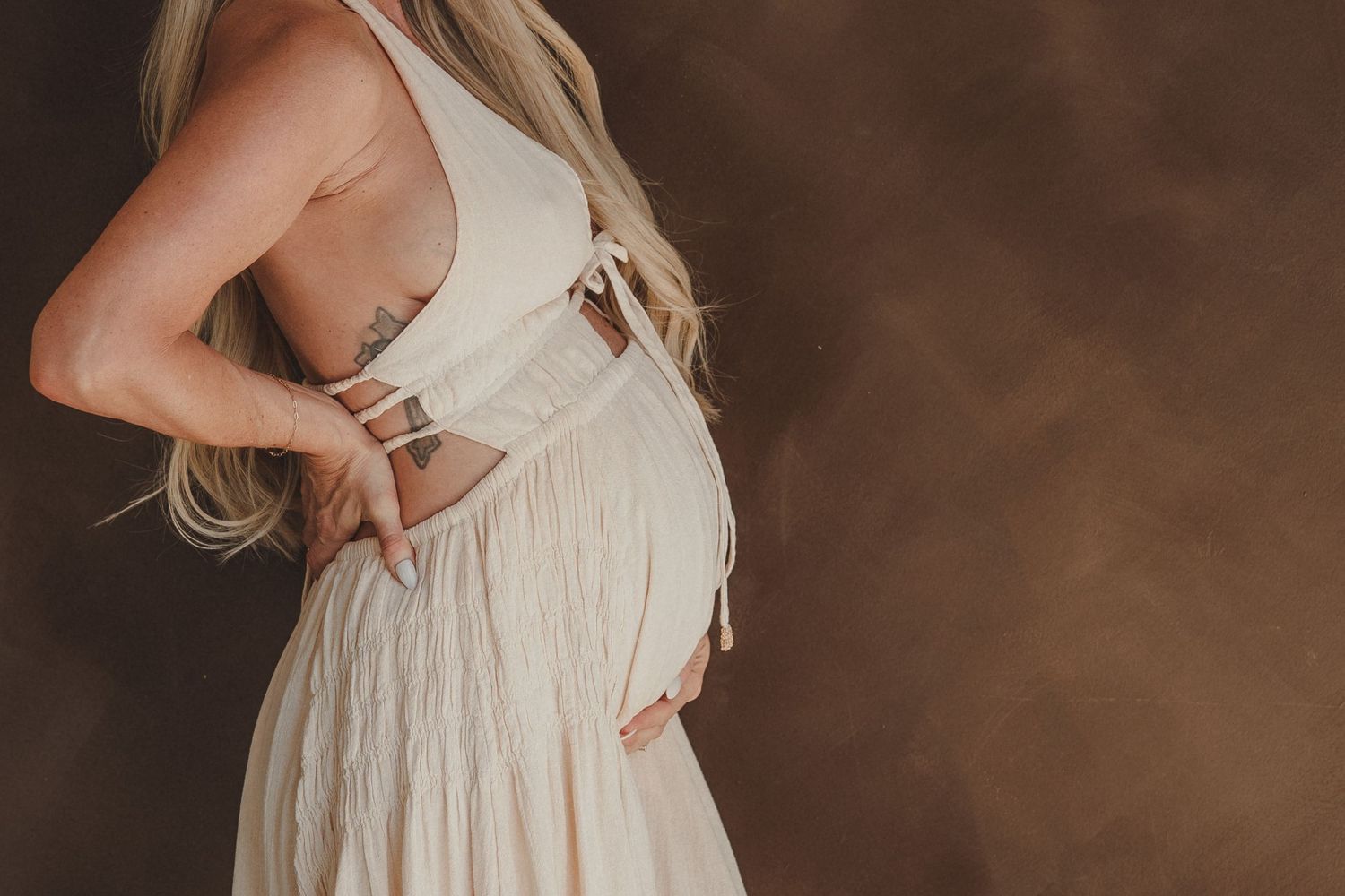 Pregnant woman in white dress cradling baby bump, side profile maternity photo with brown studio background.