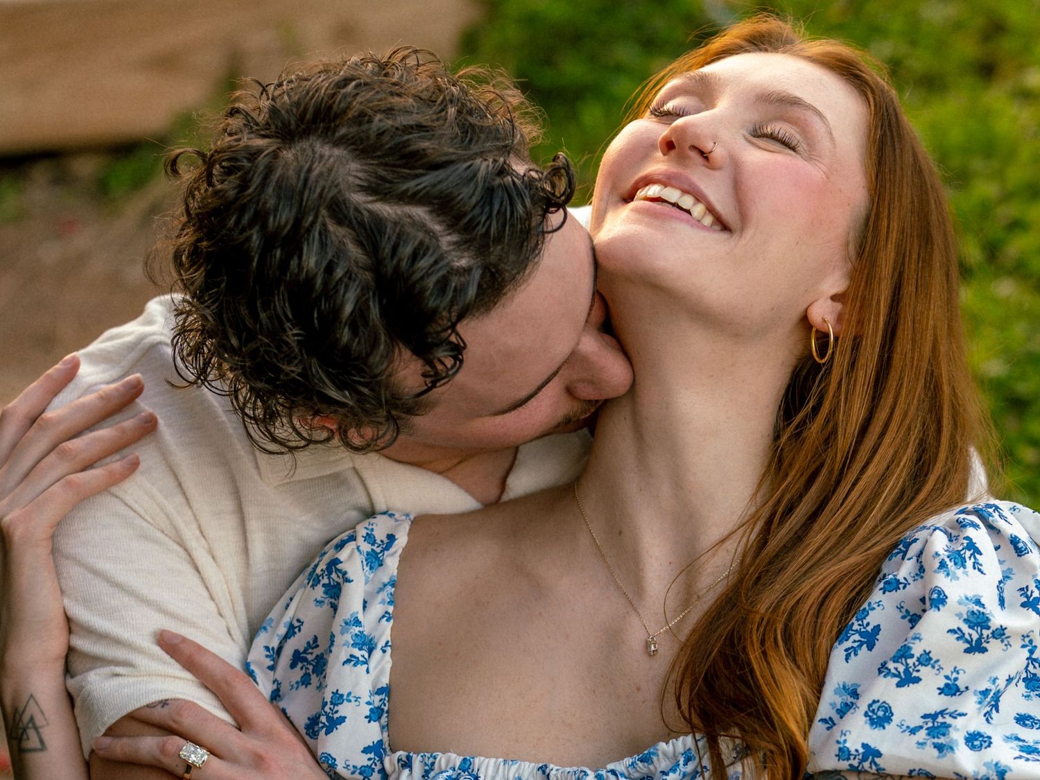 A joyful couple embracing outdoors, man kissing woman's neck as she laughs, wearing floral dress.