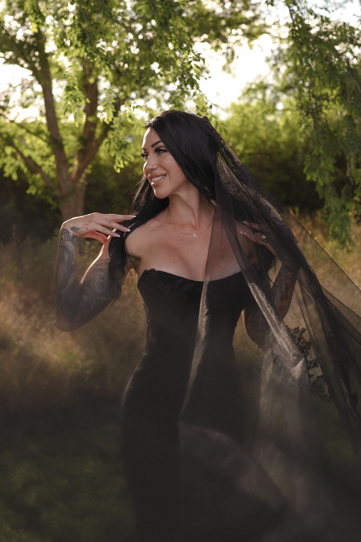Woman in black off-shoulder gown with sheer veil poses outdoors in golden hour light.