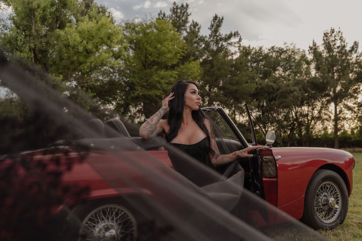 Dark-haired woman in black dress leans against a classic red convertible car outdoors.