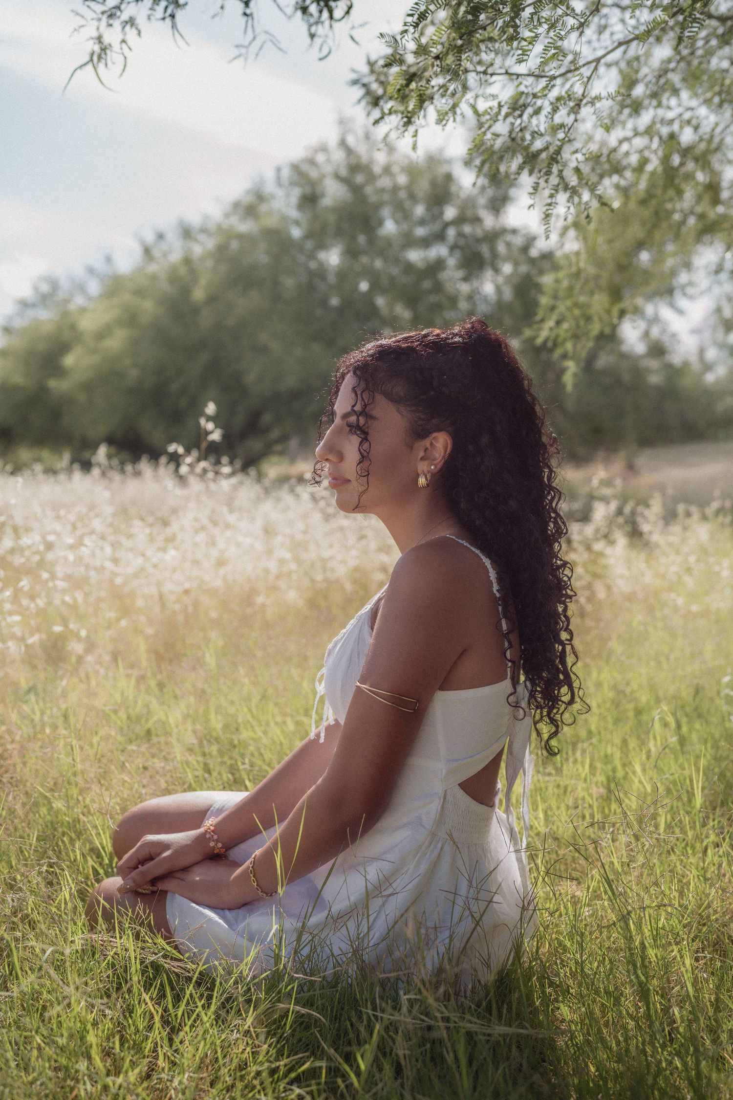 Young woman with curly hair in a white dress sitting in a sunlit meadow.
