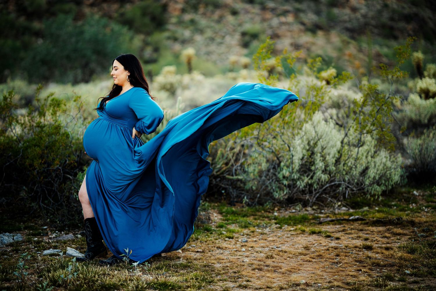 Pregnant woman in flowing teal gown poses outdoors with dress billowing in wind against desert shrub backdrop.