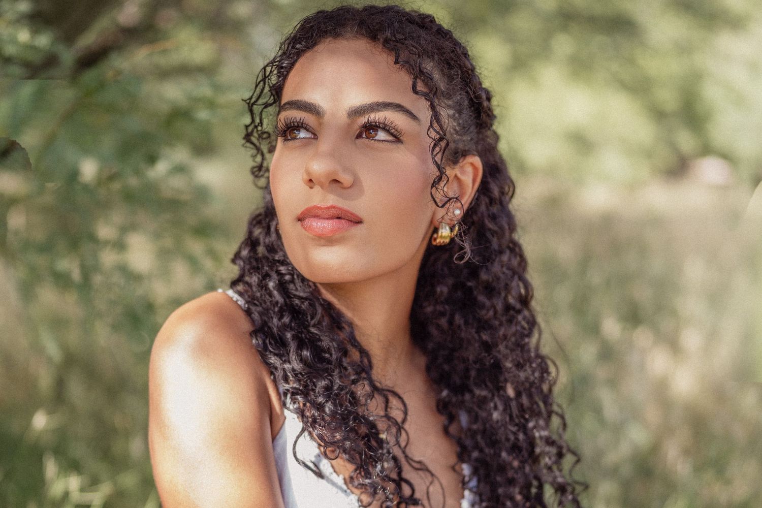 Young woman with curly hair and gold earrings poses outdoors with lush green foliage in the background.