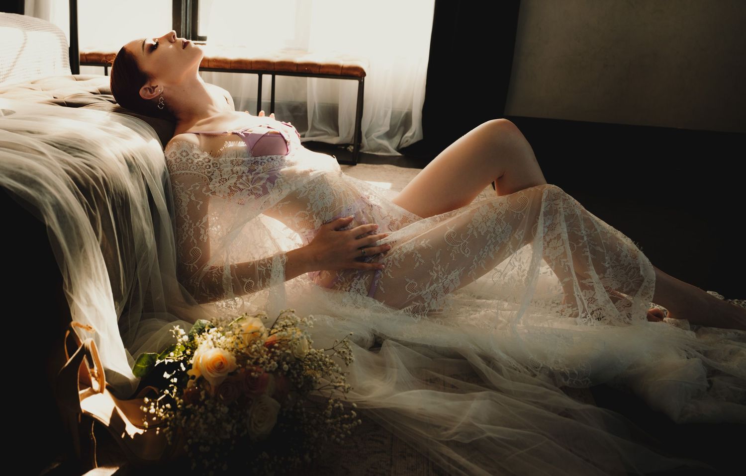 A woman in a white lace gown reclines gracefully on a sofa with soft sunlight and a floral bouquet nearby.