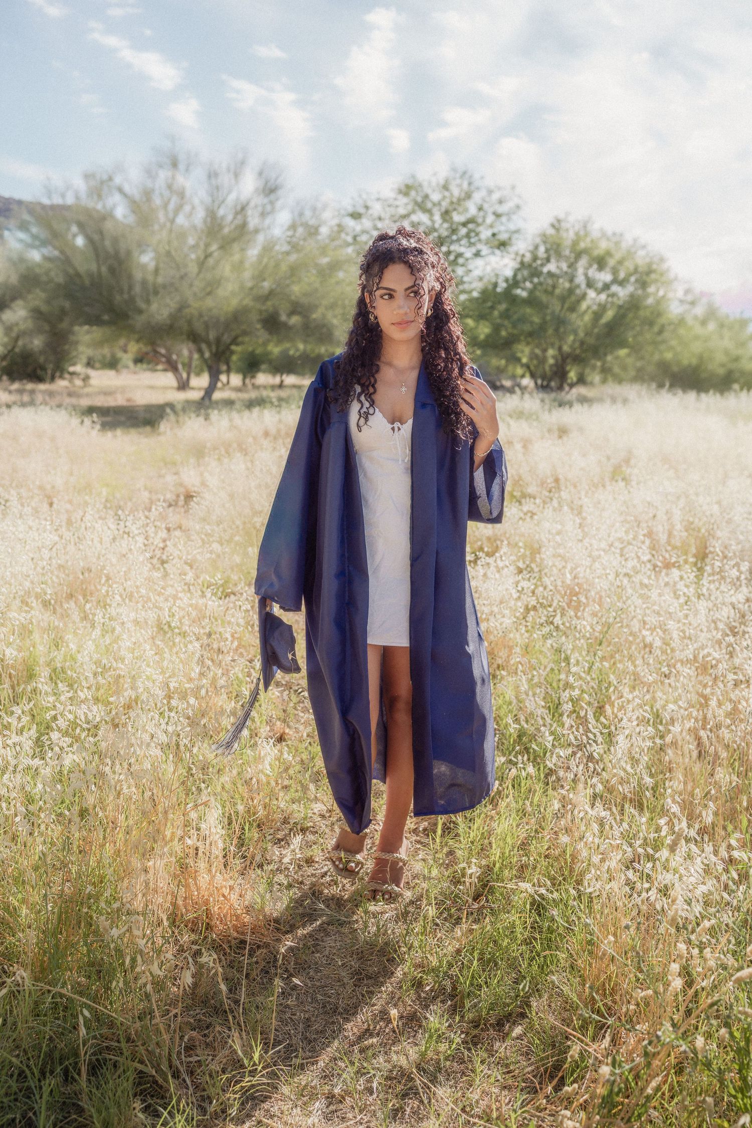 Graduate in blue robe standing in golden meadow with trees during outdoor photo session.