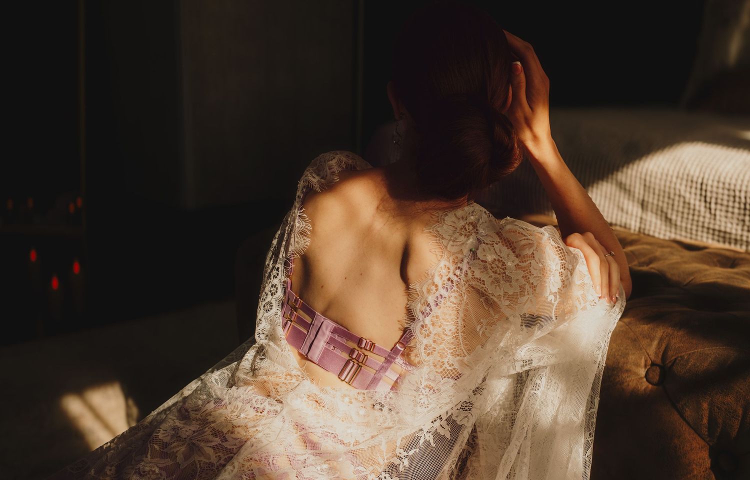 Woman in white lace robe sitting in warm sunlight, back exposed, hand in red hair, moody boudoir photography.