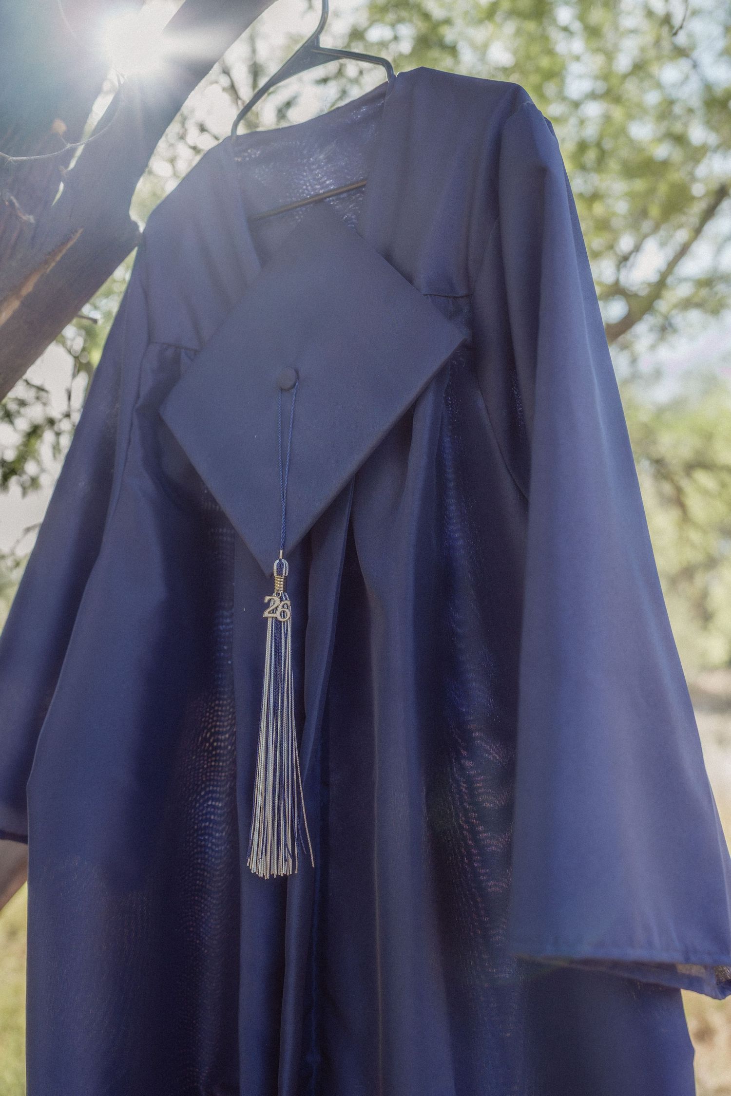 Navy blue graduation cap and gown hanging on a tree hanger outdoors with sunlight filtering through green leaves.