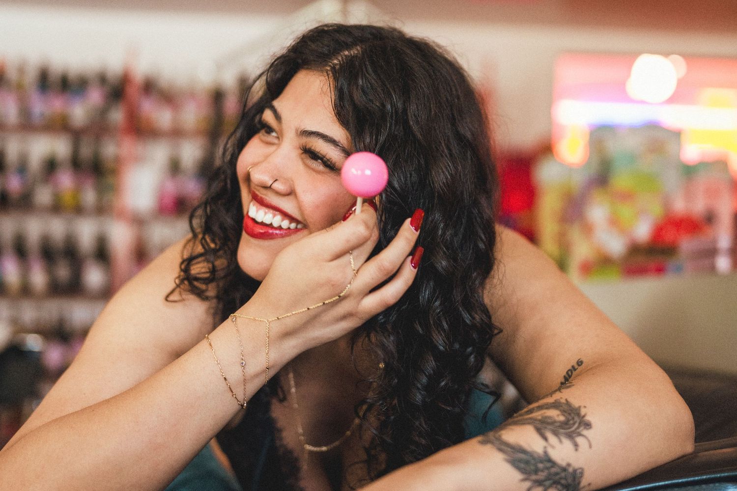 Smiling woman with curly dark hair holds a pink ball near her face in a colorful salon setting.