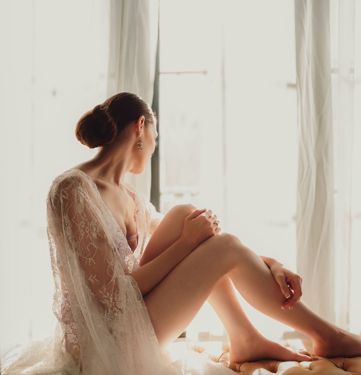 Elegant woman in lace robe sitting by a sunlit window in a serene, dreamy boudoir setting.