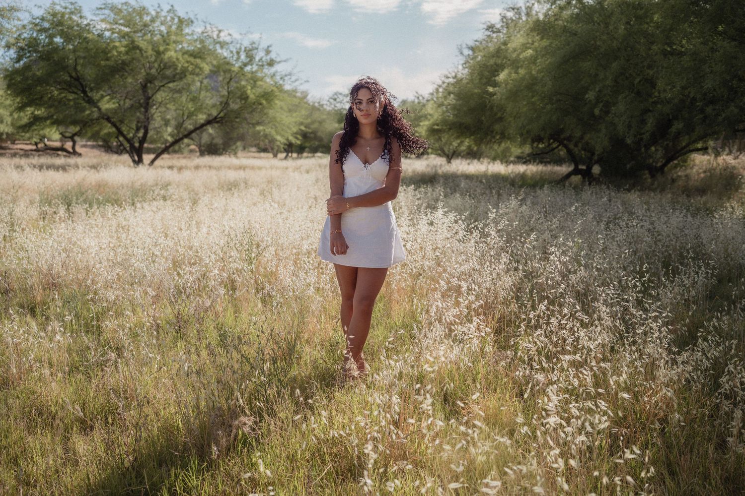 Young woman in white dress standing in a sunlit meadow with tall grass and trees in the background.