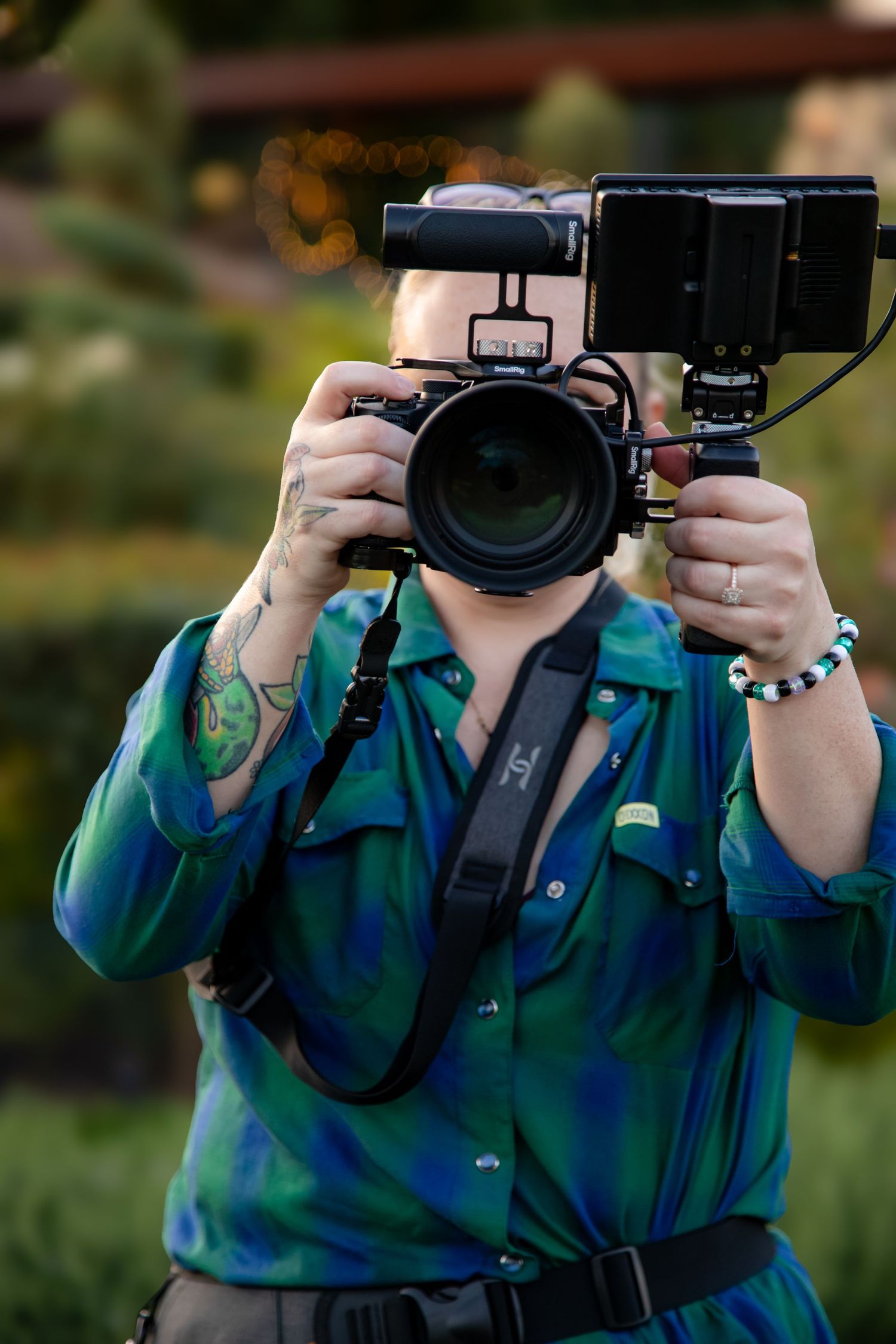 Tattooed photographer in teal shirt holds professional camera with rig up to face outdoors.