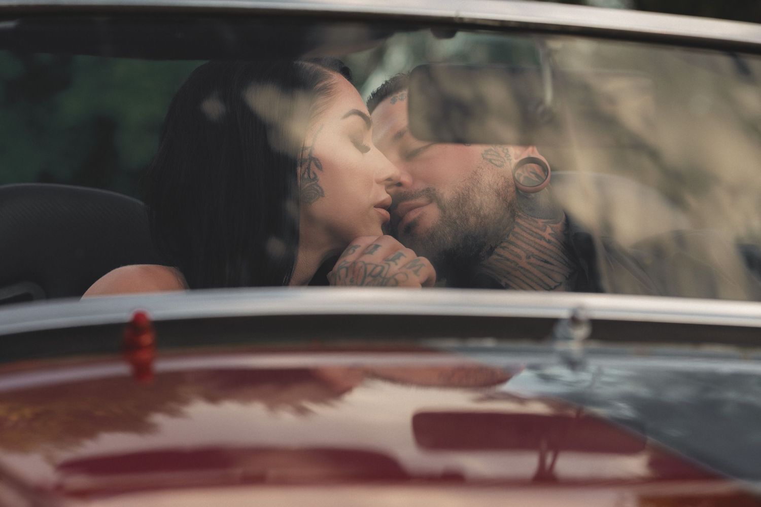Couple sharing a romantic kiss inside a vintage red car, captured through the windshield.