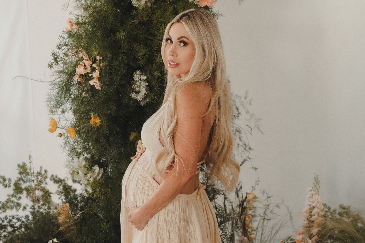 Pregnant woman in beige backless dress with long blonde hair standing before lush floral backdrop.