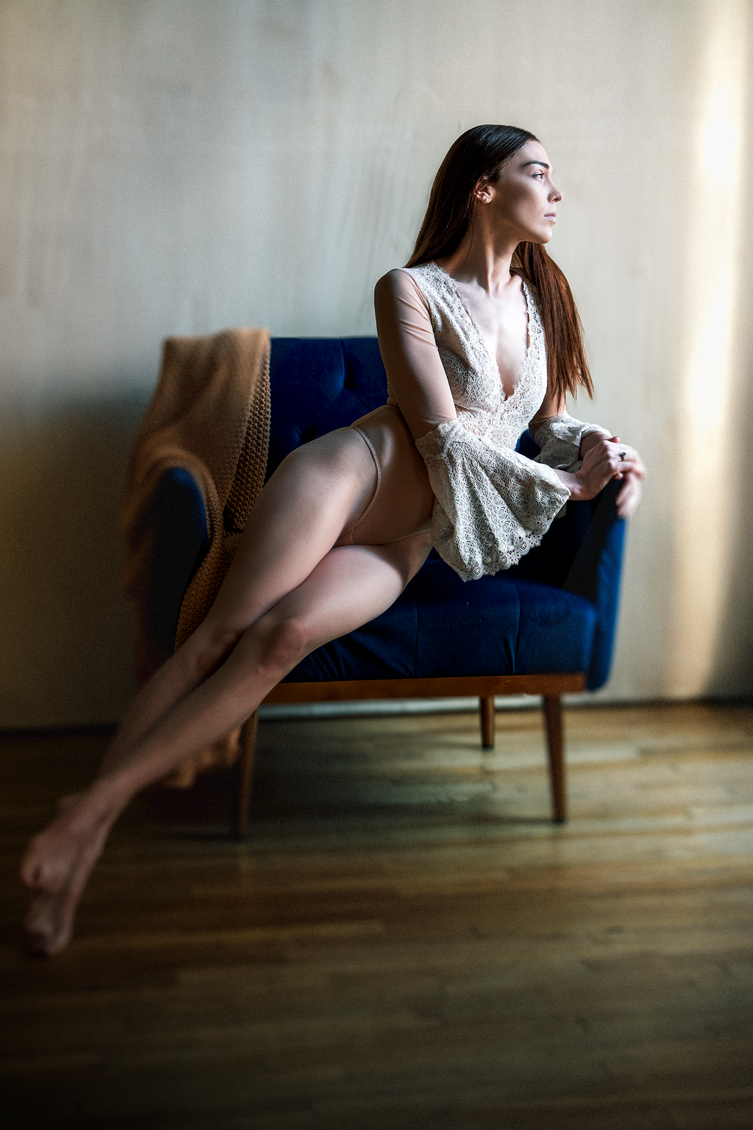 A young woman in white lace lingerie sits gracefully on a blue velvet armchair on a hardwood floor.
