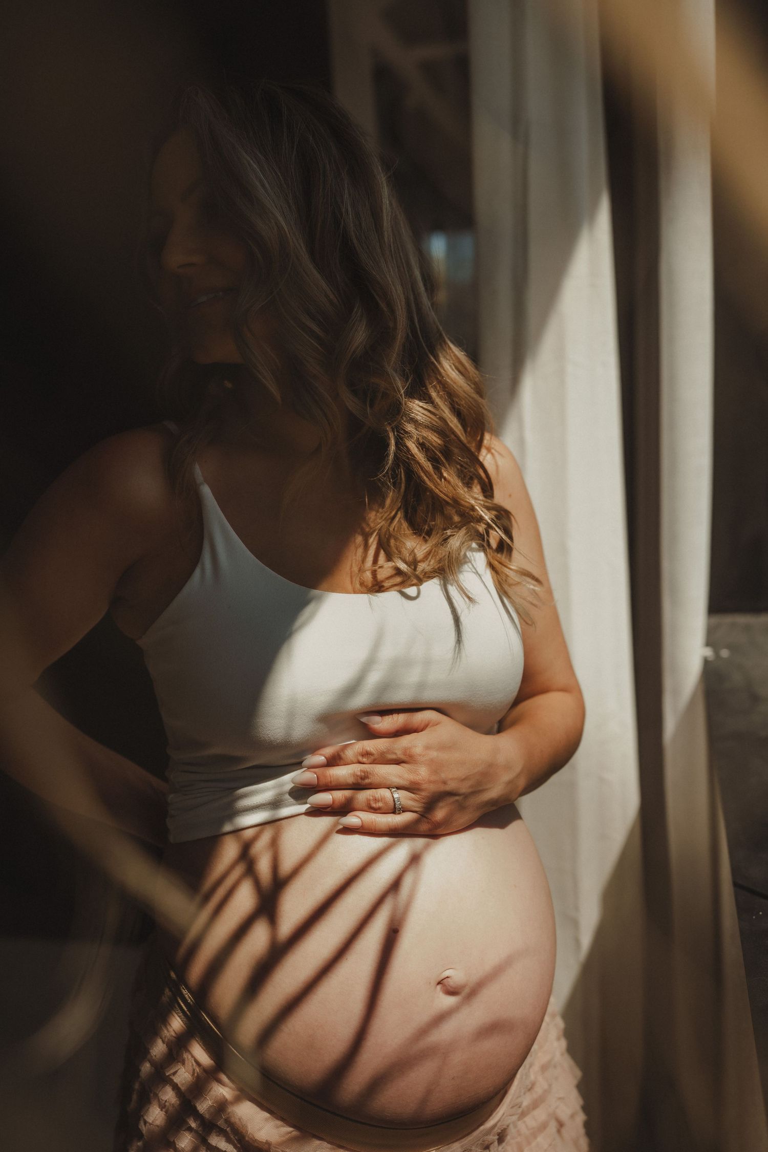 Pregnant woman in white crop top cradling her baby bump by a sunlit window with beautiful shadow patterns.