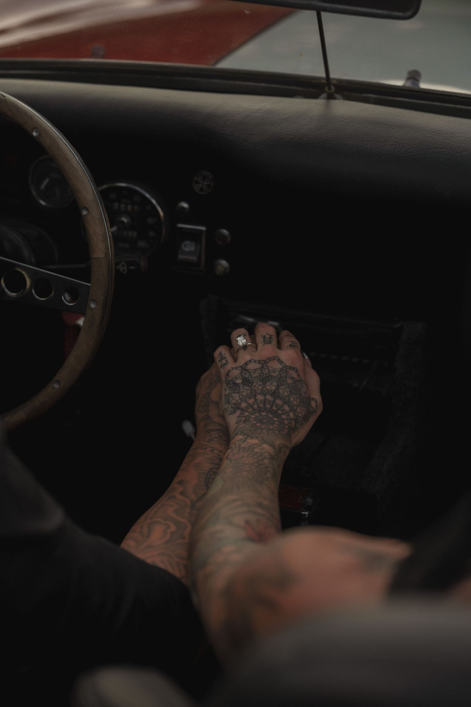 Tattooed hands resting on a vintage car dashboard near the gear shift, showcasing intricate ink designs.