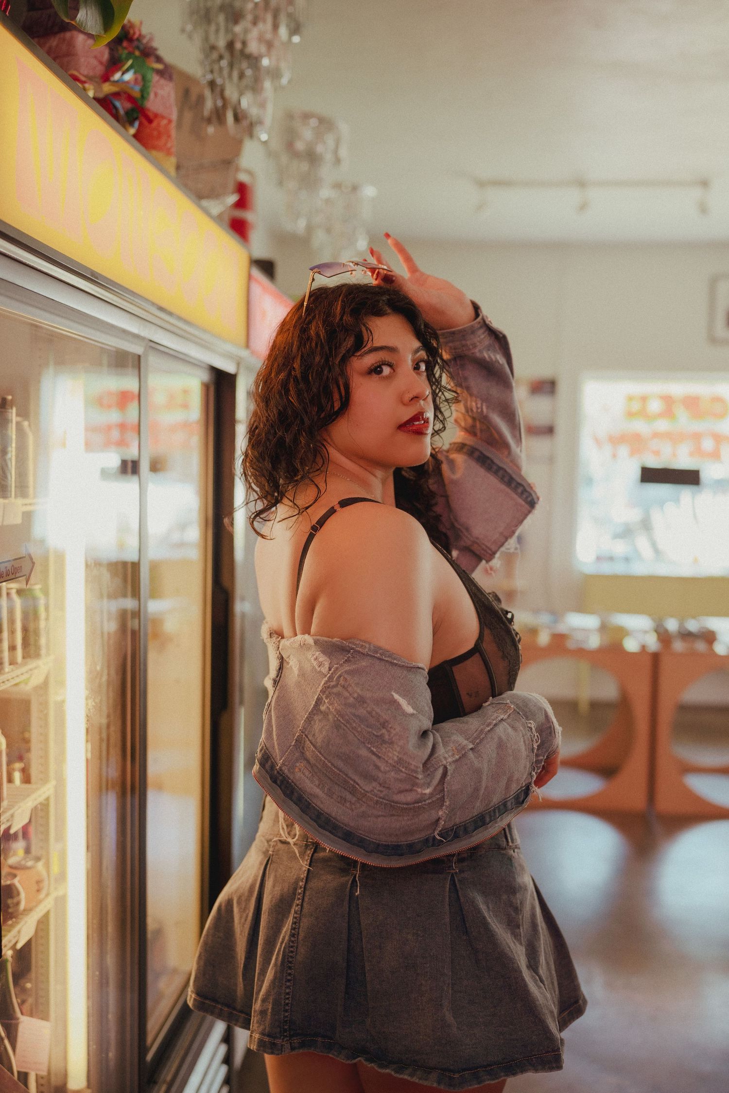 Young woman in denim jacket and skirt poses by refrigerator display in a warmly lit convenience store.