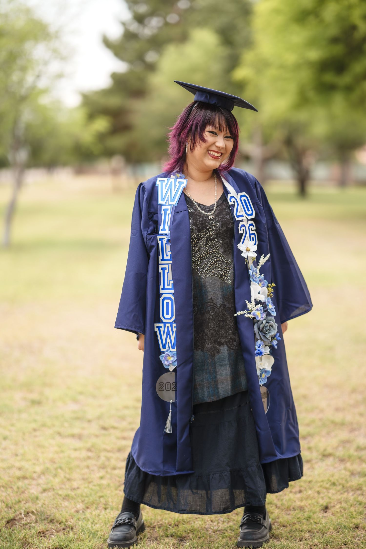 Graduate in navy blue cap and gown with personalized stole poses outdoors in park setting.