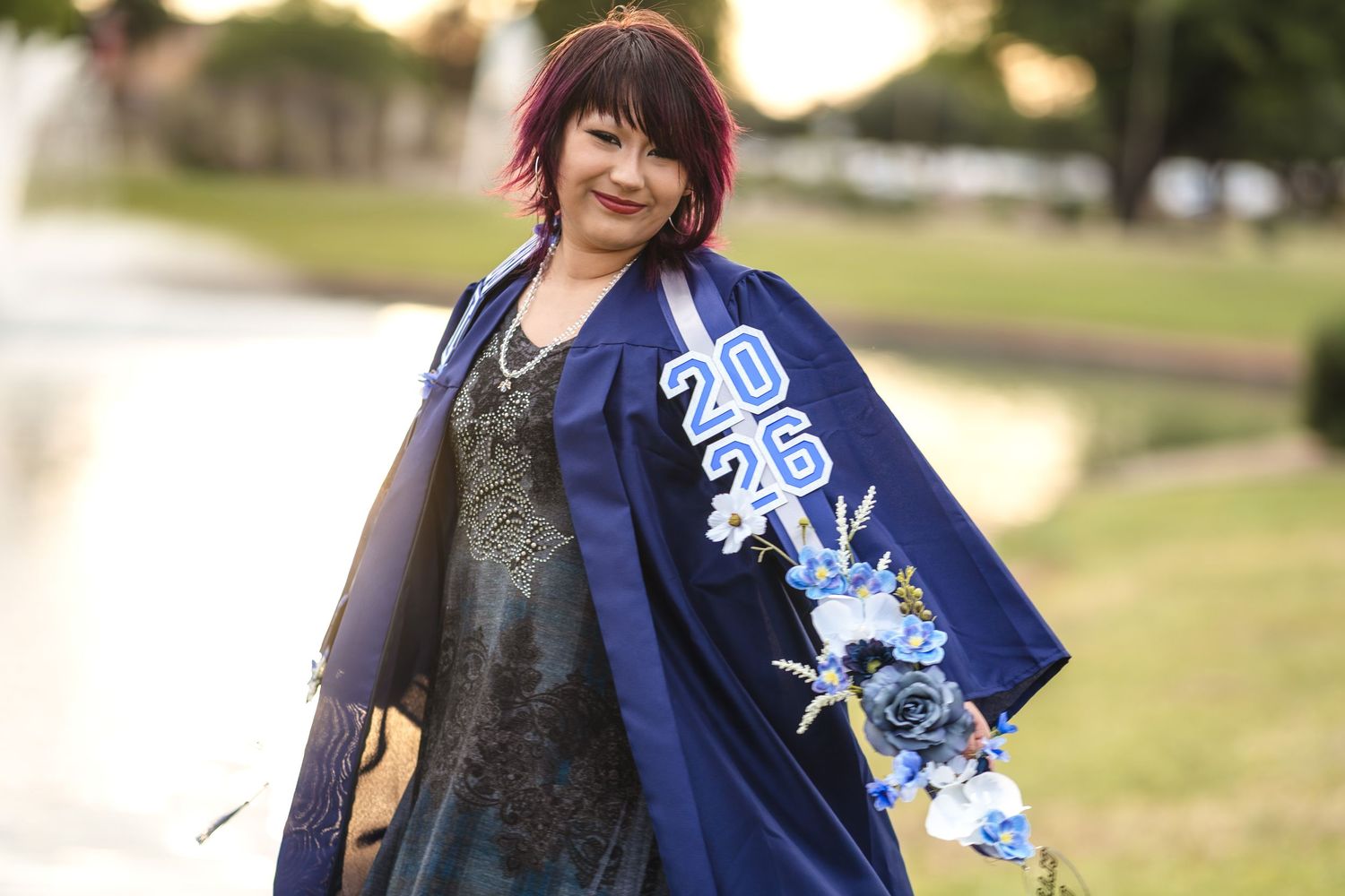 Woman in blue graduation gown with 2026 embroidery and floral design smiles outdoors near a fountain.