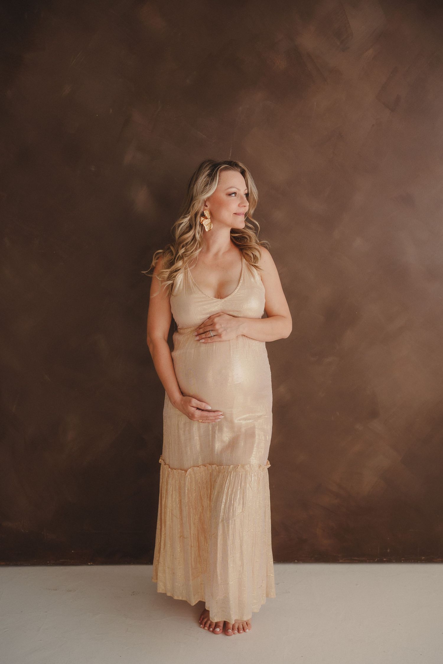 Pregnant woman in sheer beige maxi dress holding baby bump against brown textured backdrop.
