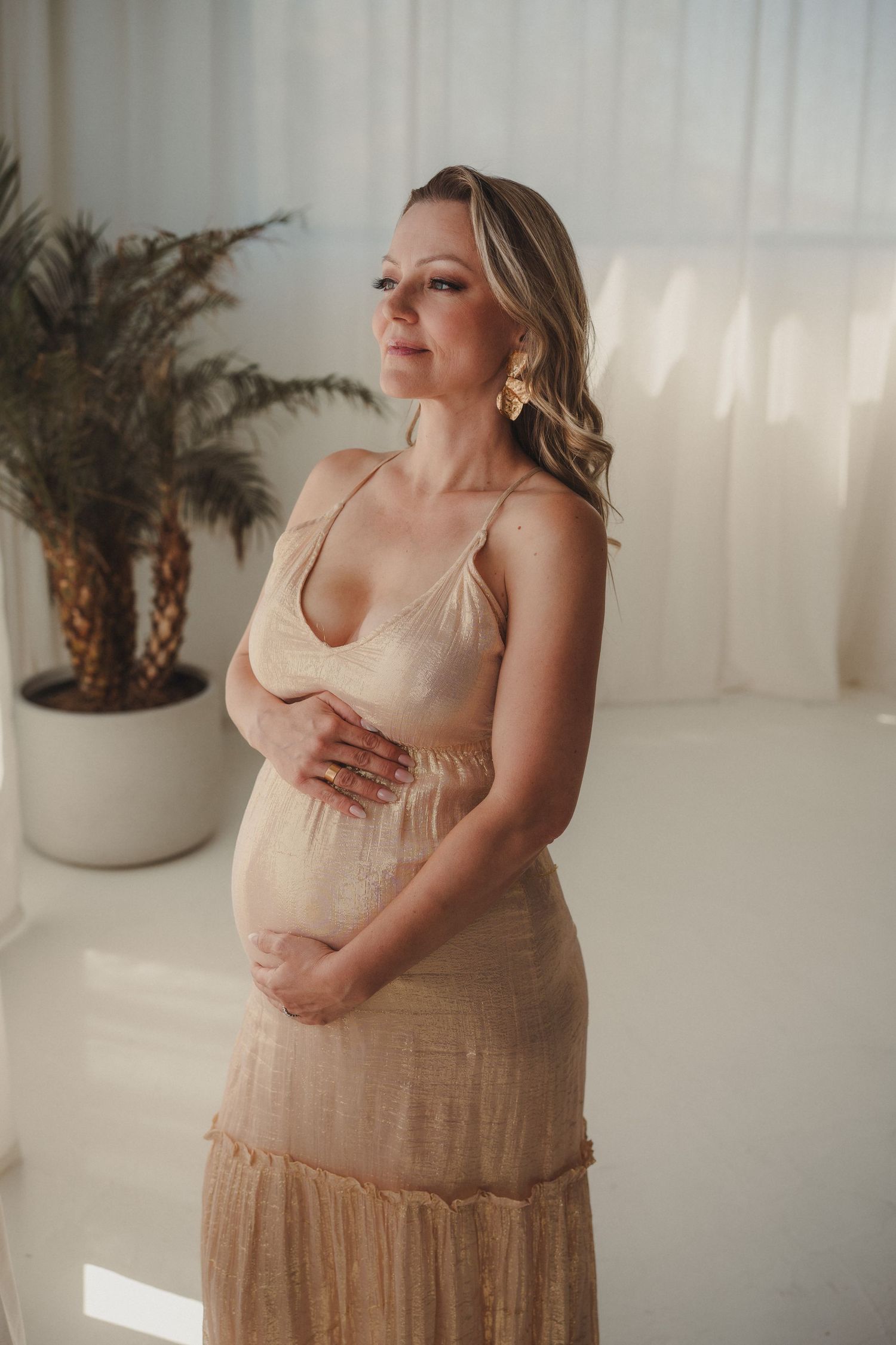 Elegant pregnant woman in beige dress cradles her bump in a bright, airy room.