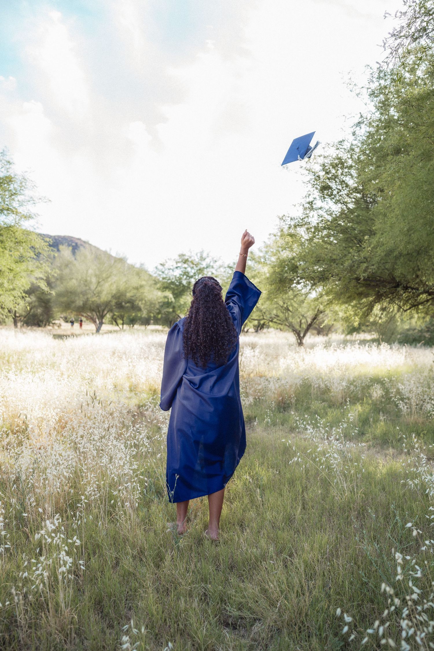 Graduate in blue gown tossing cap in sunny meadow surrounded by green trees and wildflowers, celebrating achievement outdoors.