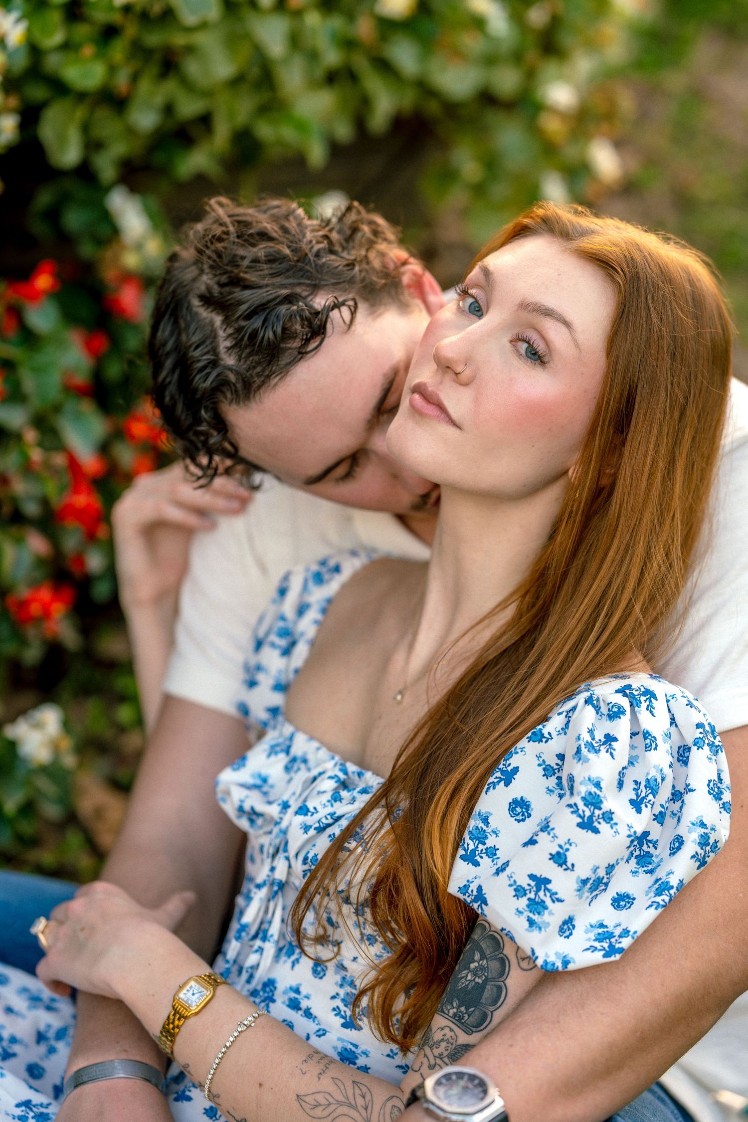 A couple shares a romantic moment outdoors, man kissing redhead woman's cheek near blooming flowers.