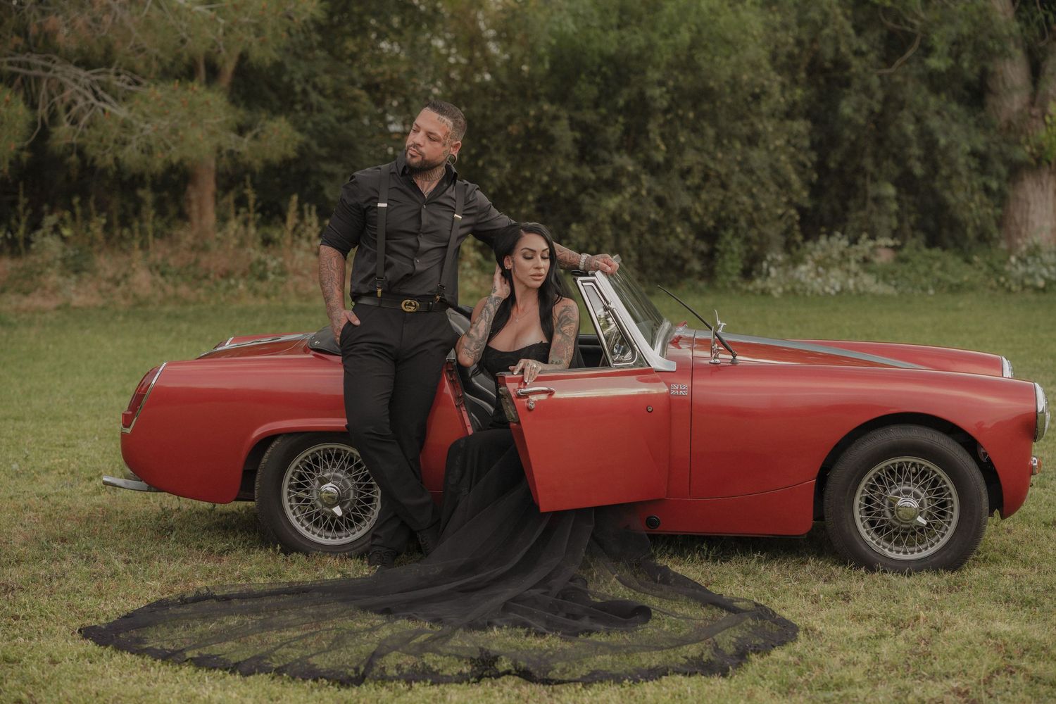 Couple posing with a classic red convertible sports car on green grass, woman in black gown, man in black outfit.