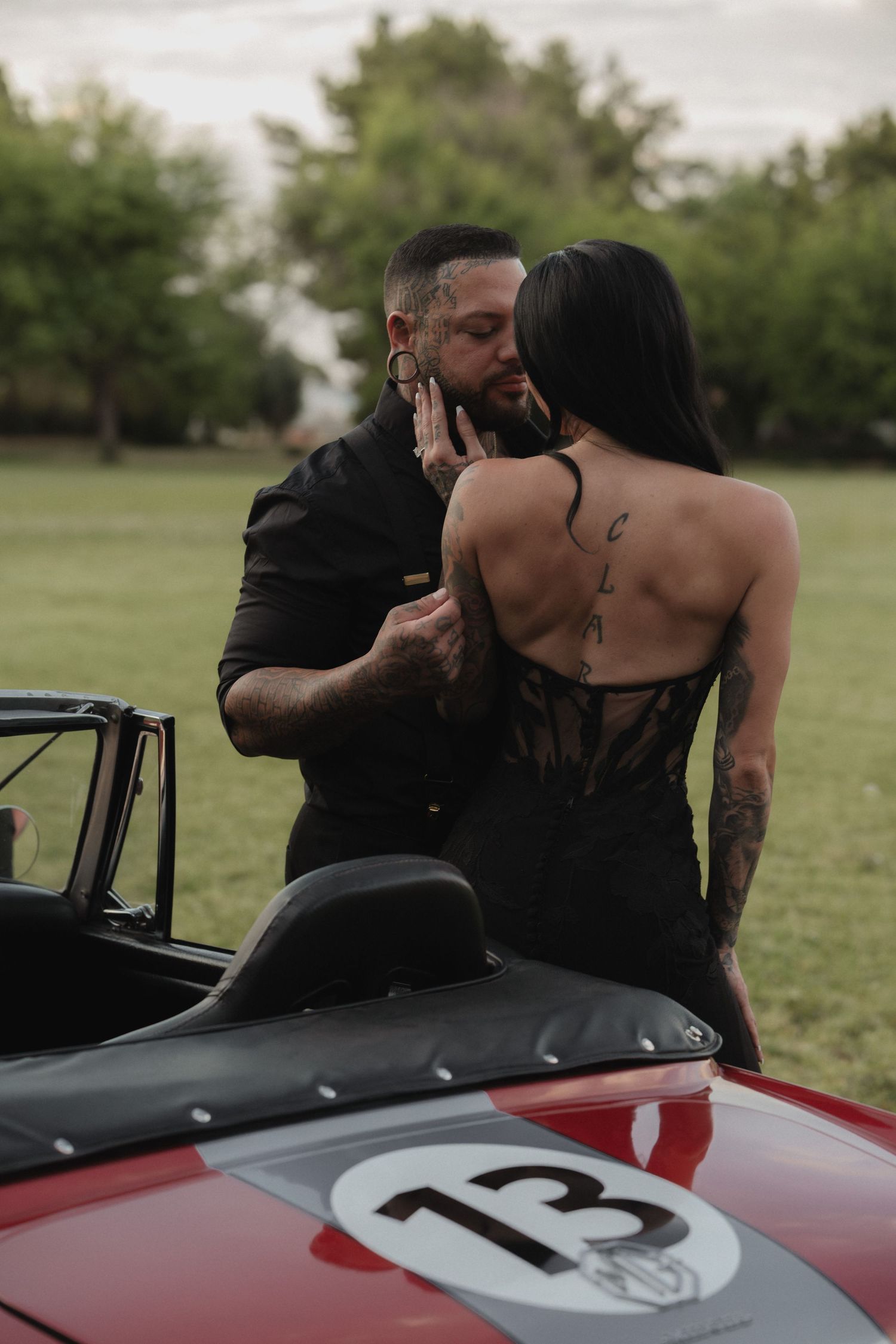 Tattooed couple sharing a romantic kiss beside a vintage red race car numbered 13 in an outdoor setting.