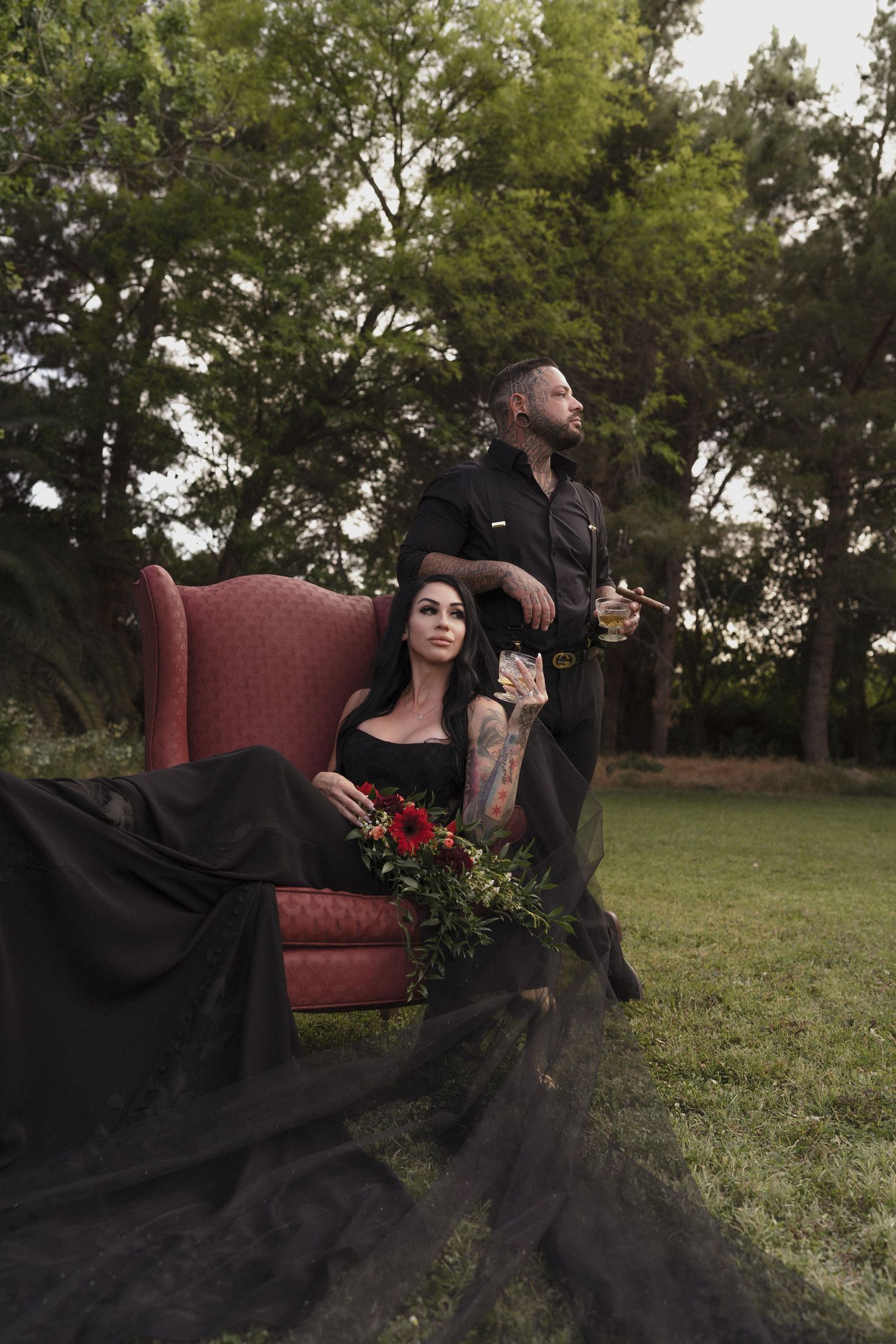 Tattooed couple poses in garden with red velvet chair, woman in black gown holding red flowers.