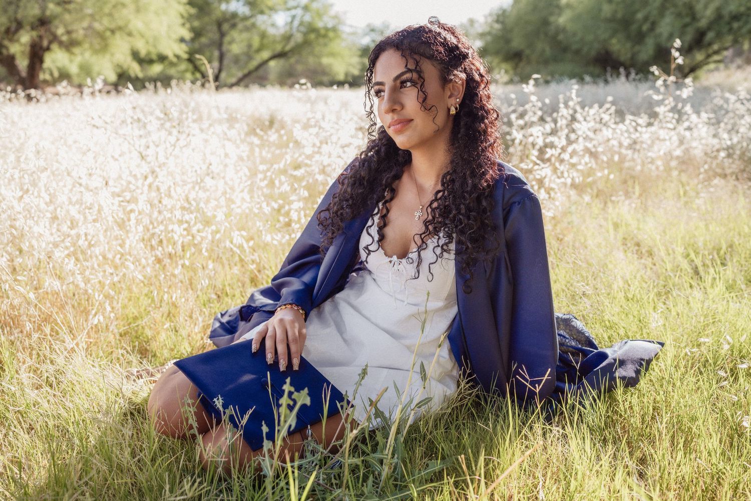 Woman with curly hair sitting in a grassy field wearing a blue jacket and white dress, gazing thoughtfully.