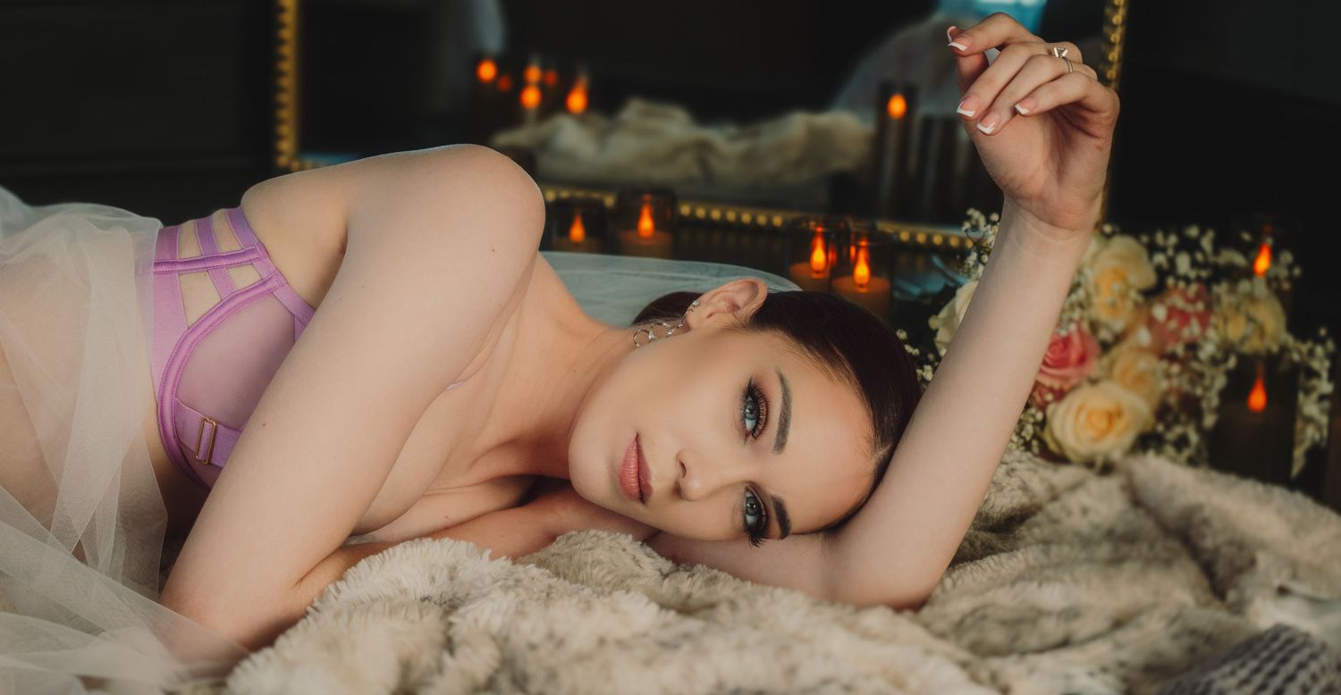 Young woman in lavender lingerie lying on a fur blanket with candles and flowers in a moody boudoir setting.