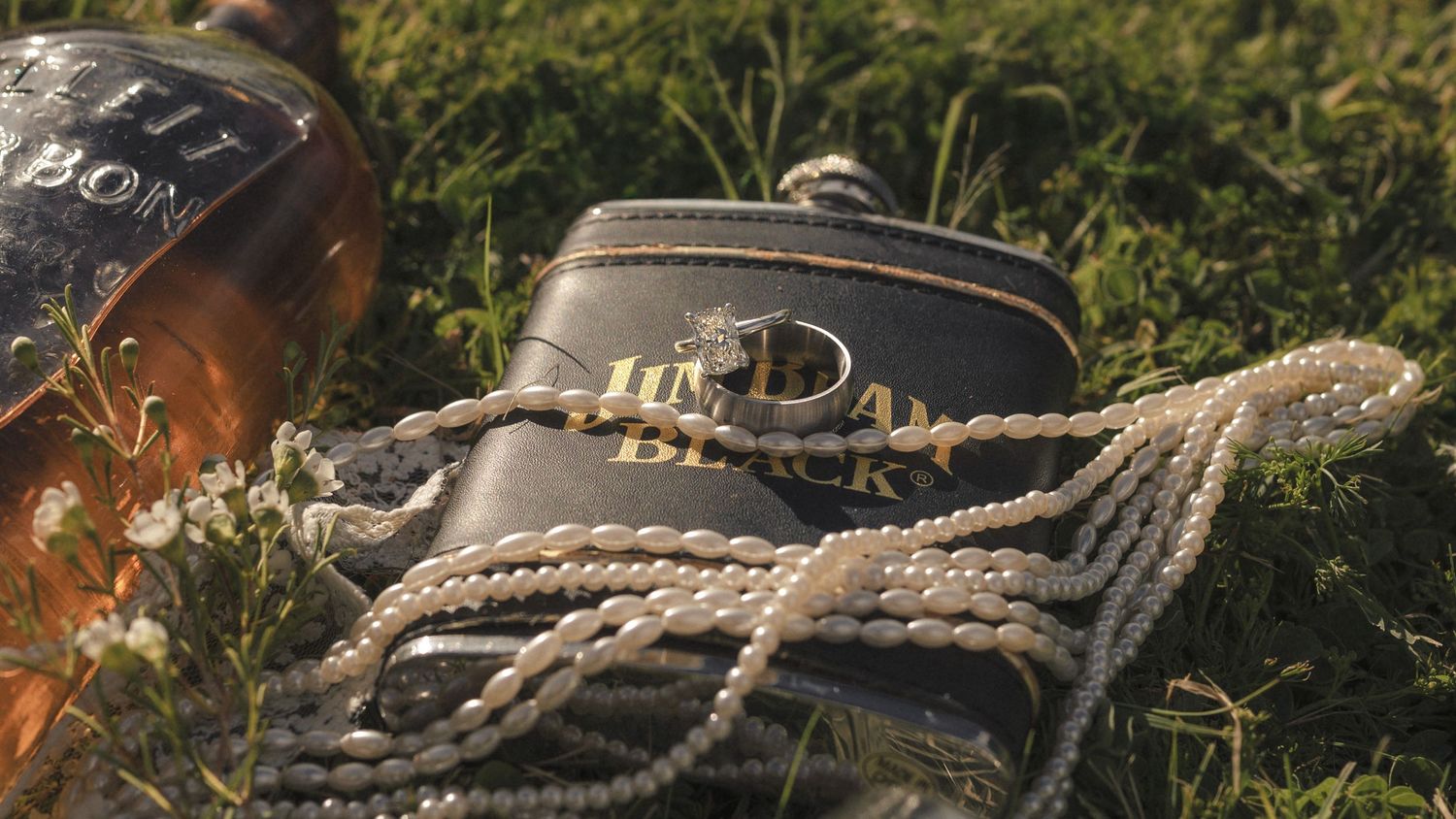 Close-up of leather wallet with wedding rings and pearl necklace resting on green grass.