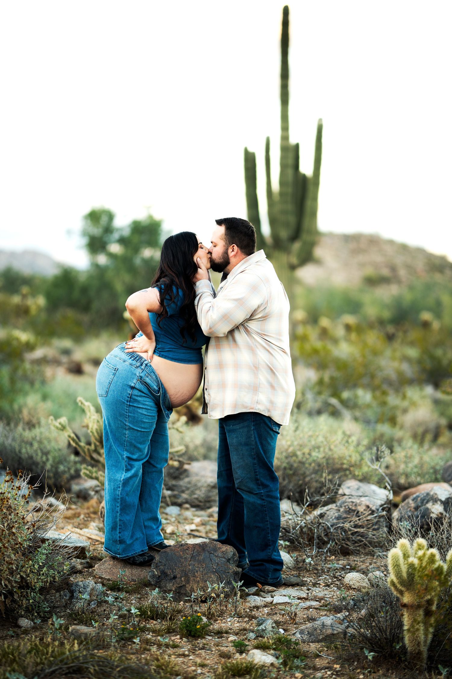 Pregnant couple sharing a tender kiss in a desert landscape with saguaro cacti, woman showing baby bump in jeans.