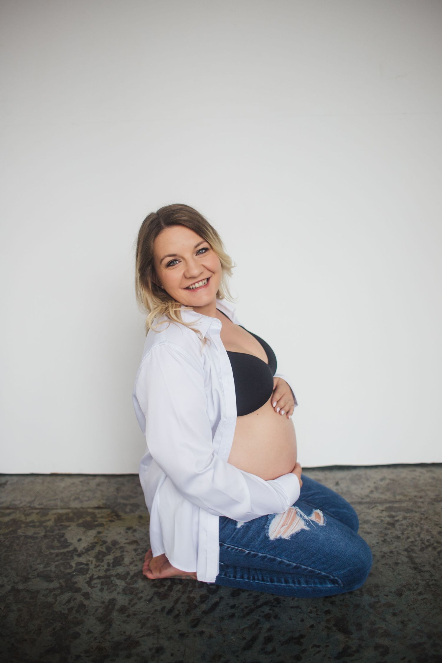 Smiling pregnant woman sitting on floor in open white shirt and jeans, cradling her bare baby bump.