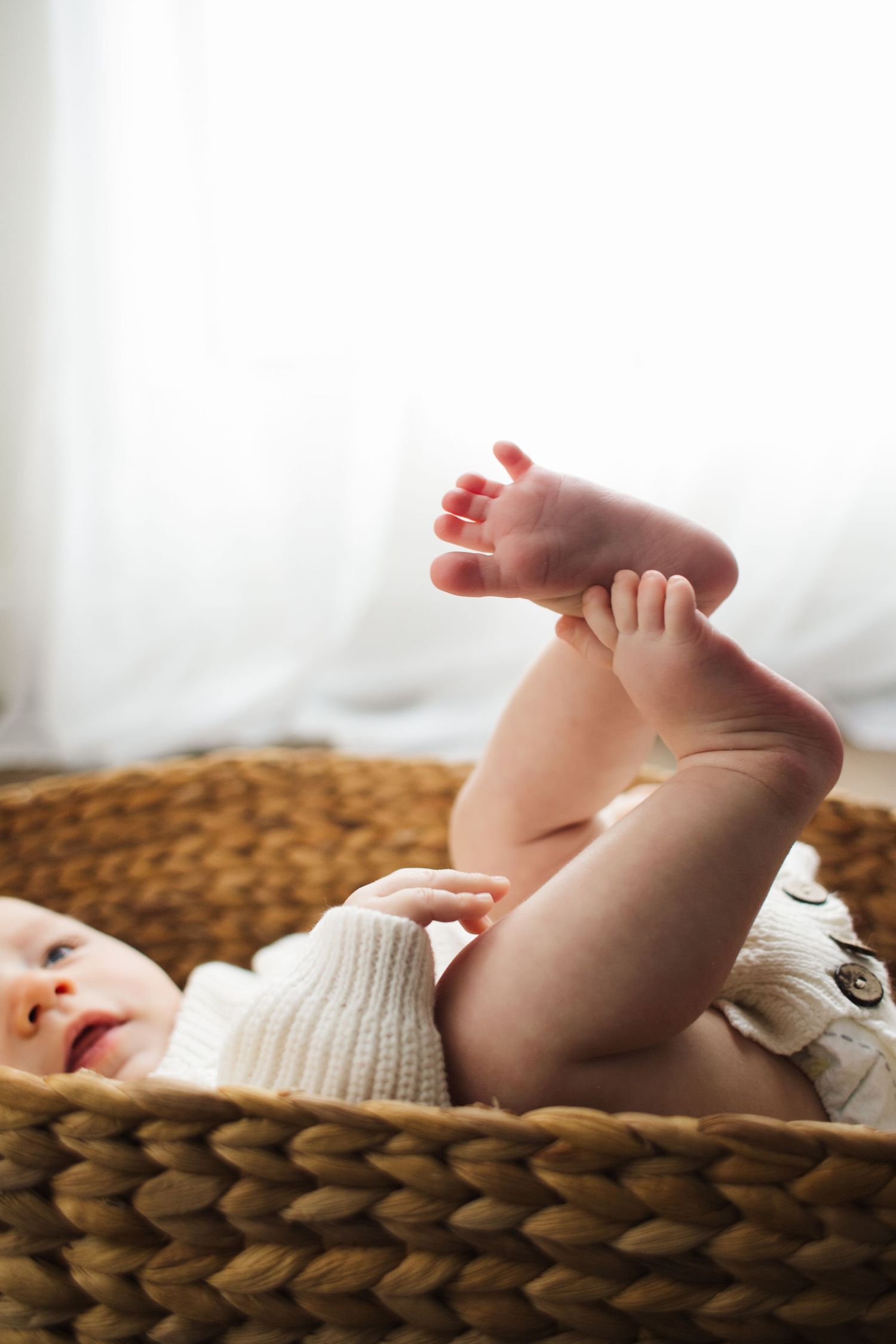 Cute baby lying in woven basket holding feet up in the air near bright window.