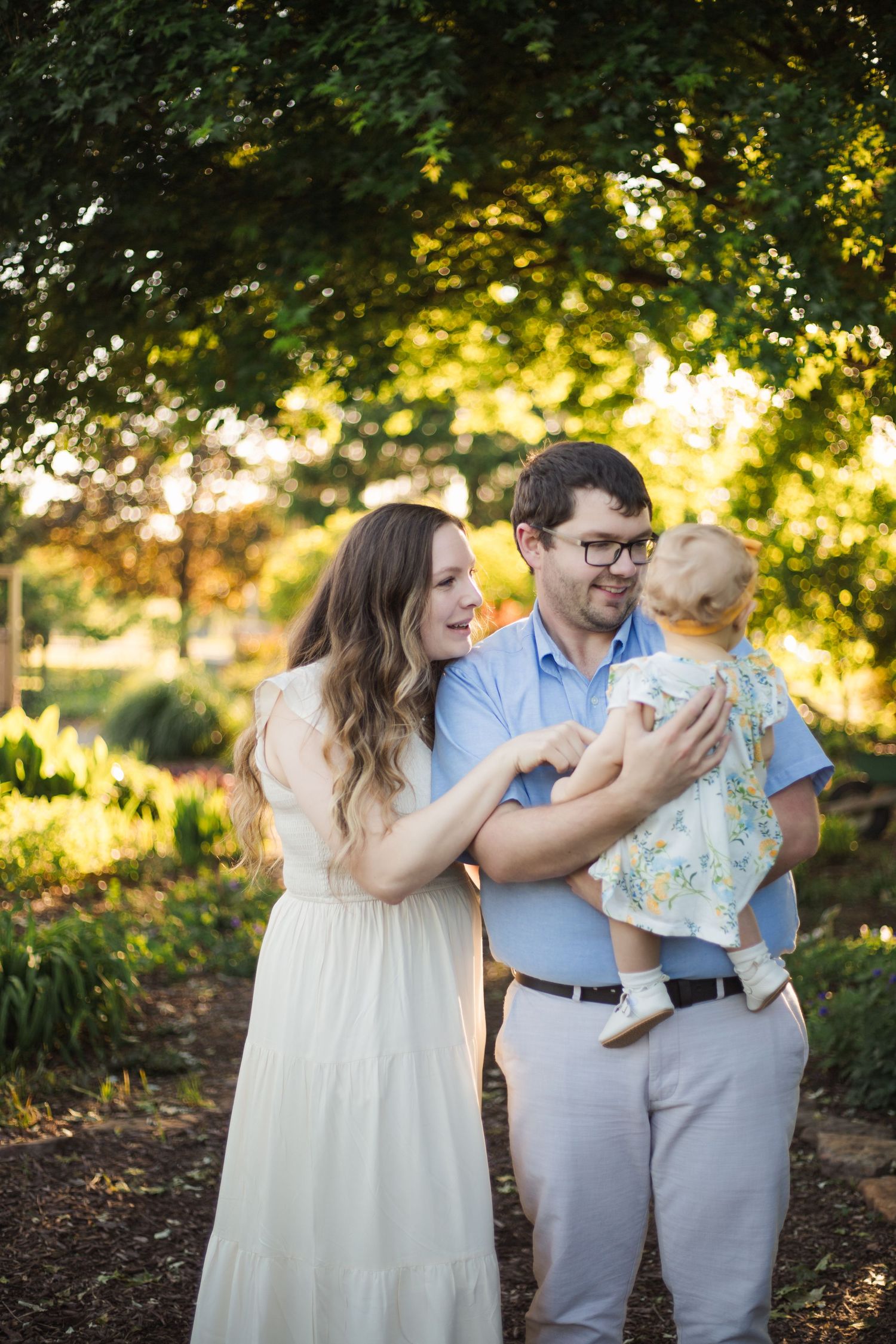 Young couple laughing together while holding their baby girl in a sun-dappled garden setting.