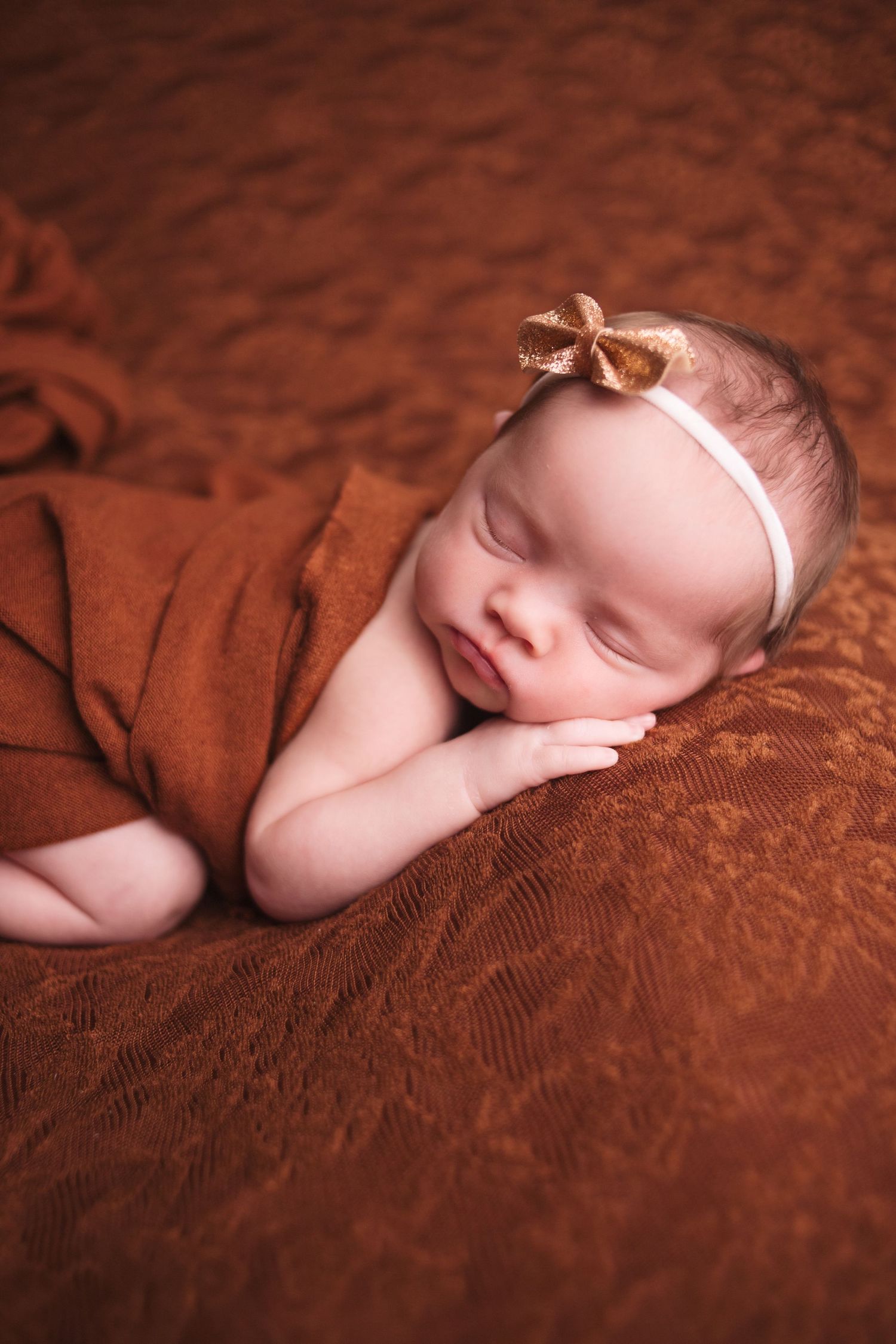 Newborn baby girl sleeping peacefully on rust brown textured blanket with gold bow headband.