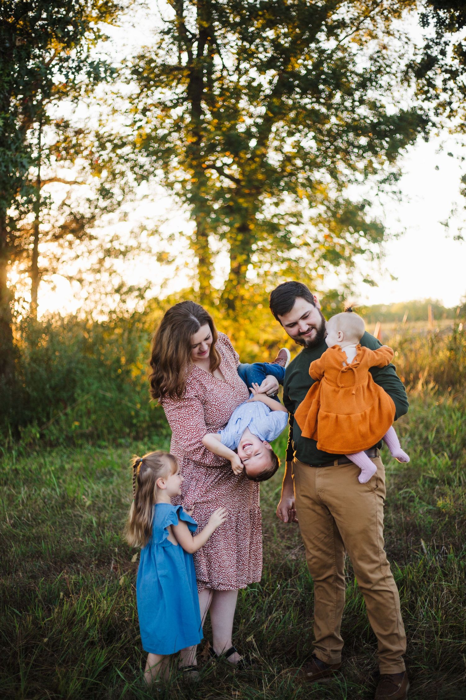 Family of five at golden hour, parents playfully holding children upside down in grassy field.