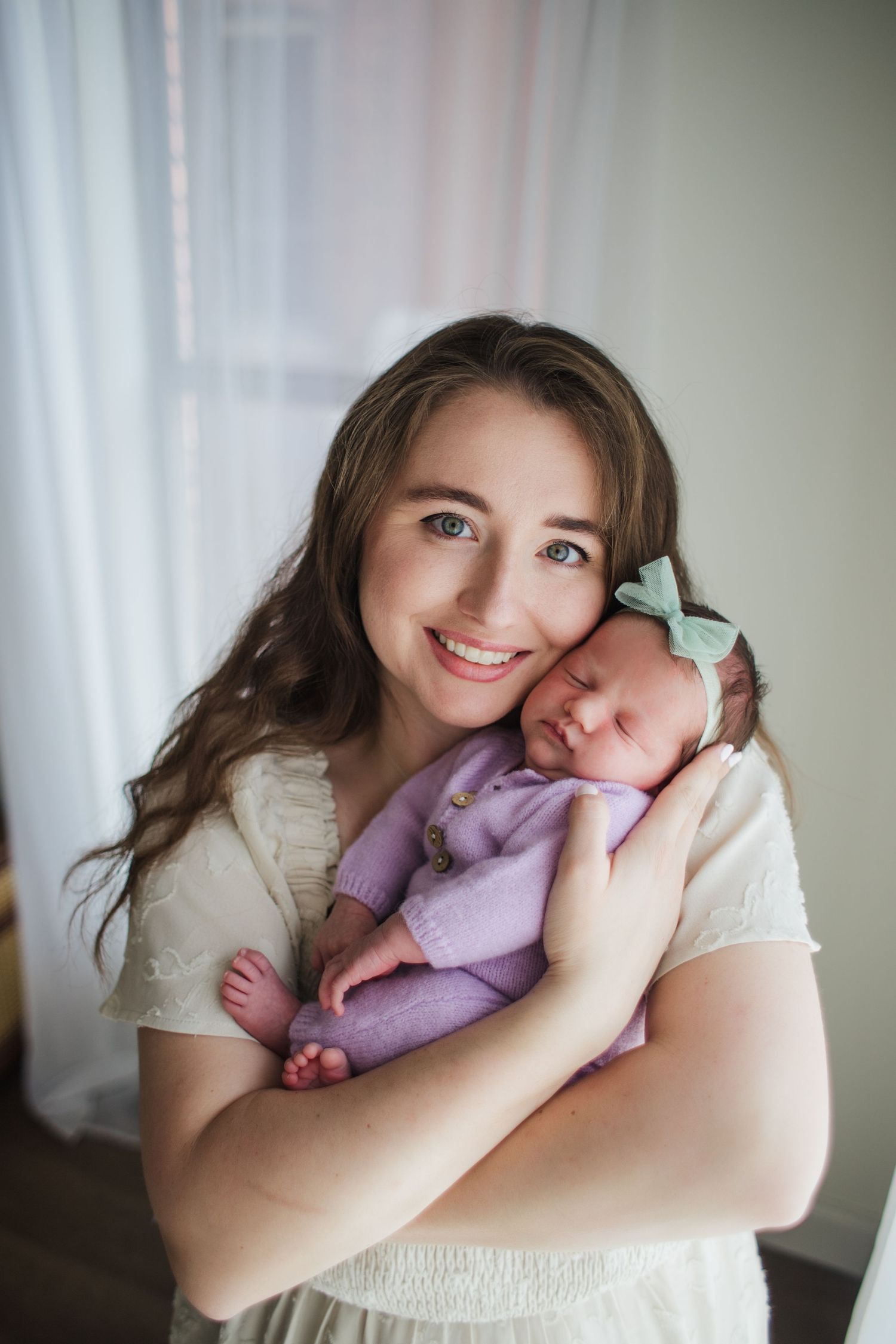 Smiling mother with blue eyes holding newborn baby in purple outfit with mint bow headband.