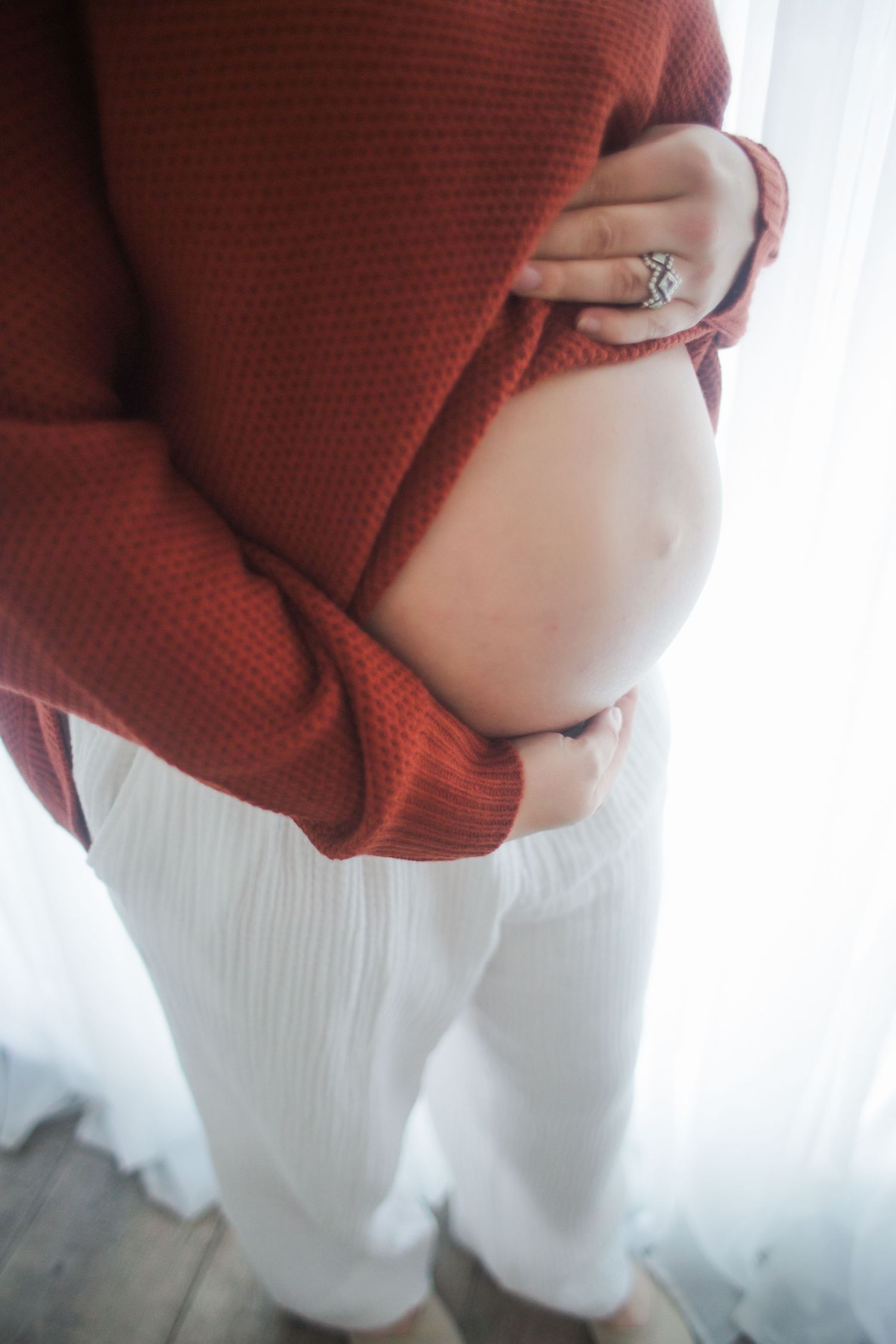Close-up of pregnant woman in rust sweater and white pants cradling baby bump.