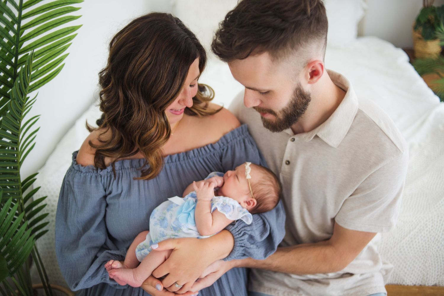 Happy parents holding their newborn baby together in a bright, plant-filled nursery room.
