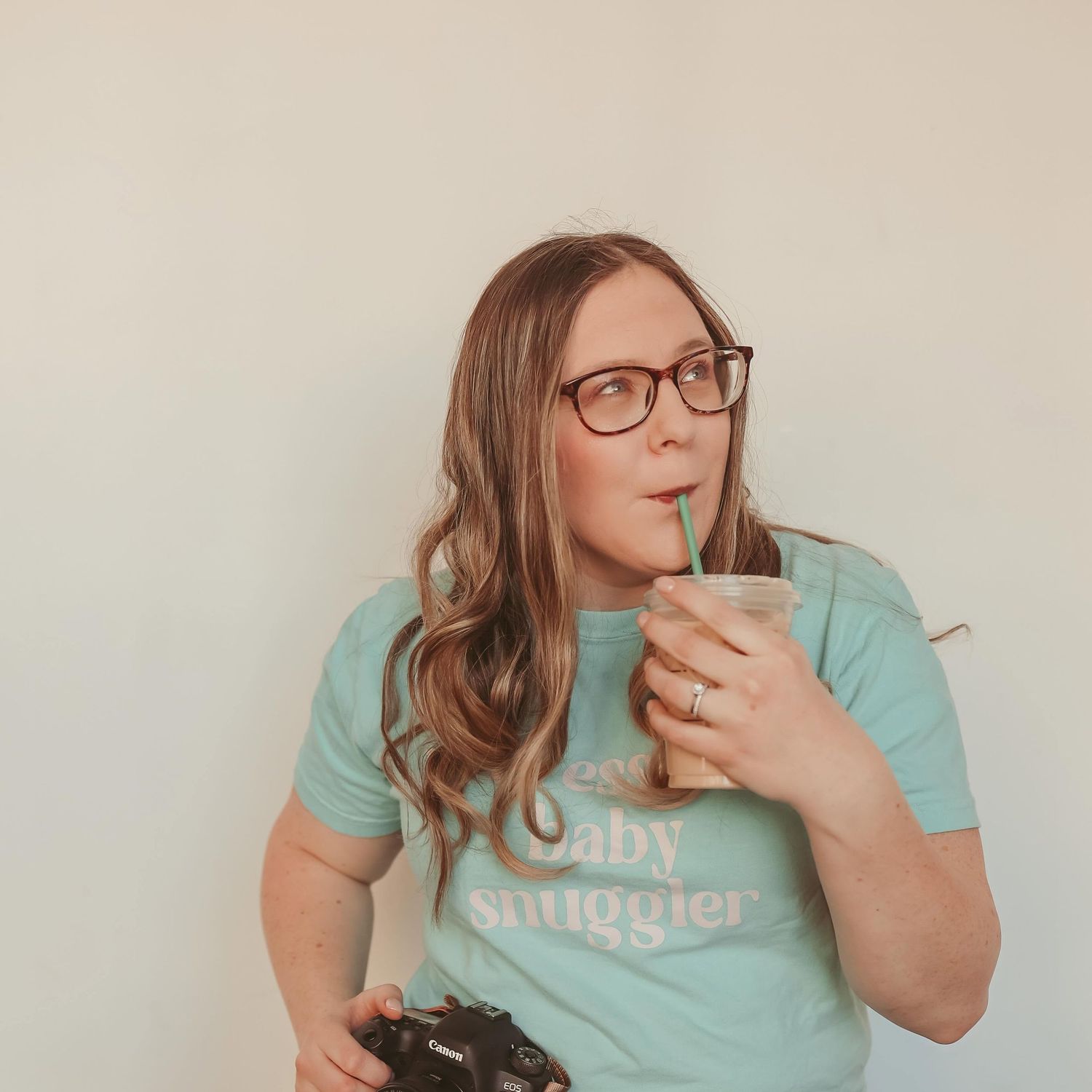 Female newborn photographer holding iced coffee drink and Canon camera wearing mint baby snuggler t-shirt.