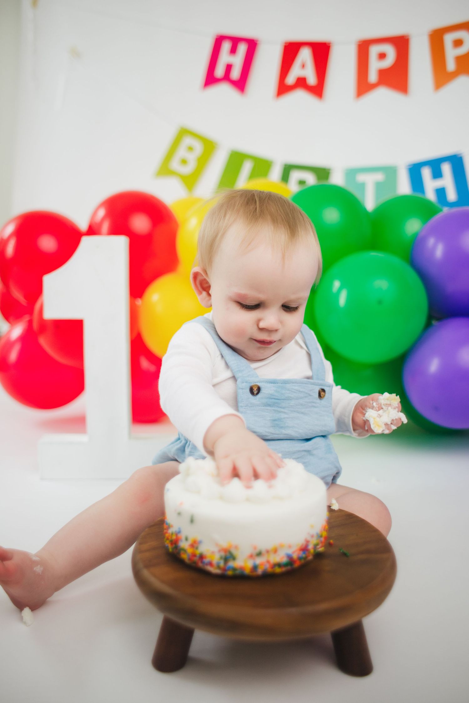 Baby celebrating first birthday smashing white sprinkle cake with colorful rainbow balloons backdrop.