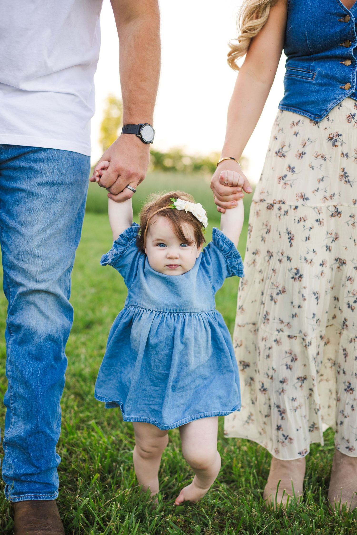 A toddler girl in a blue dress holds hands with her parents while walking barefoot on green grass.