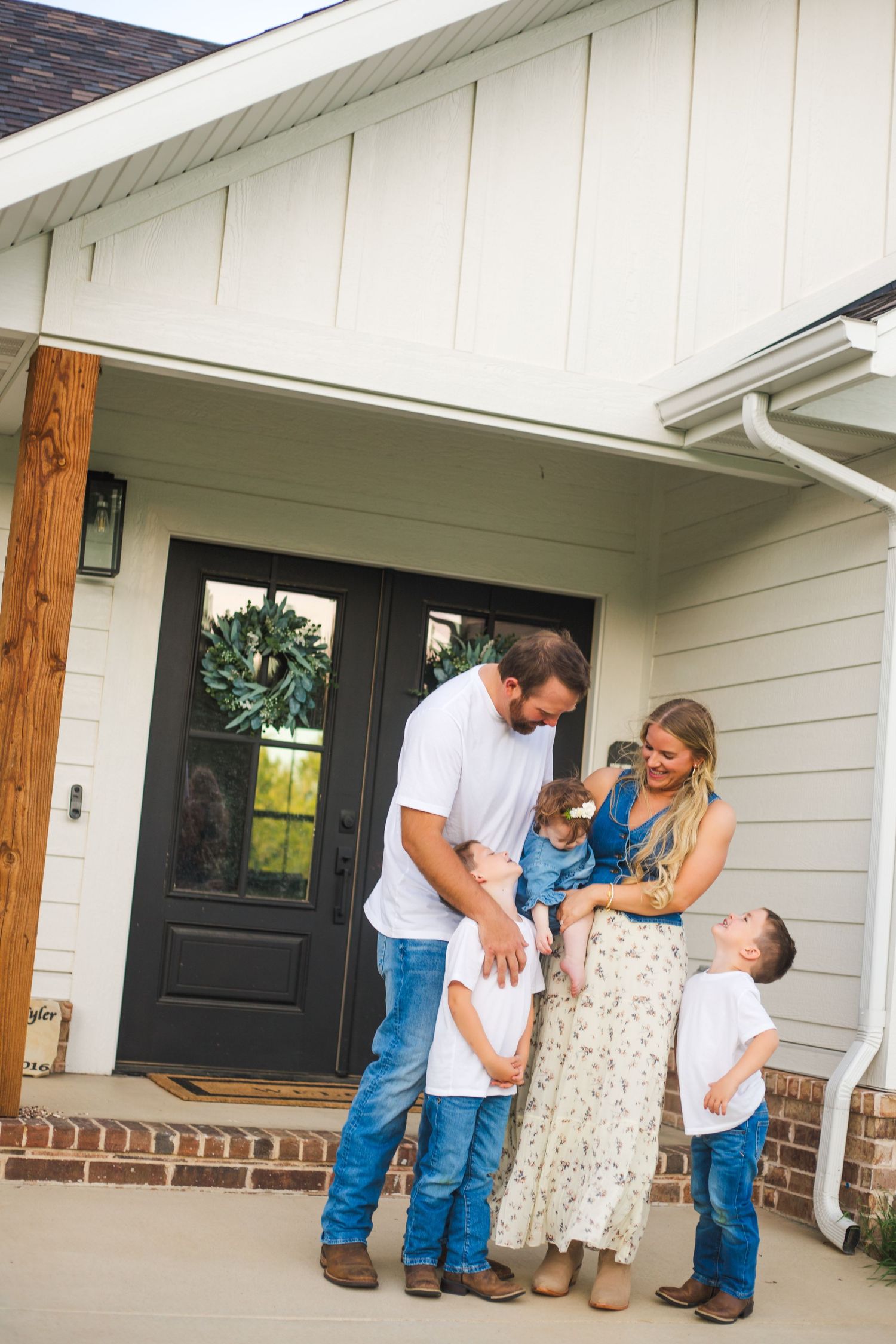 A happy family of five laughs together on the porch of a modern farmhouse home.