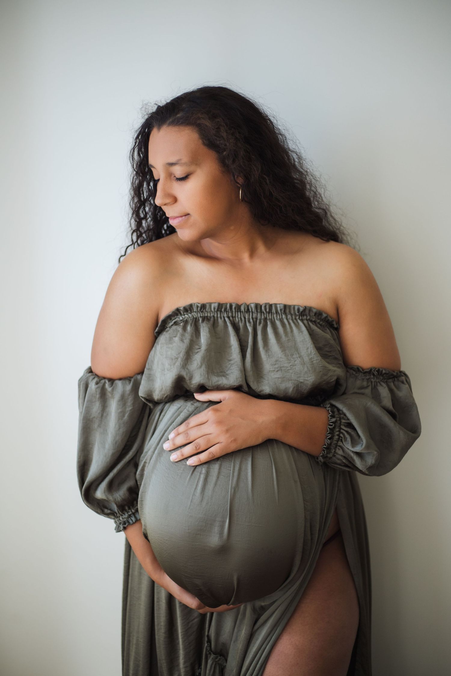 Pregnant woman in olive off-shoulder dress cradling baby bump against light background.