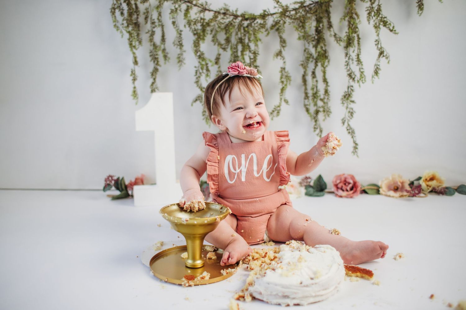 A joyful baby girl in a pink romper celebrates her first birthday smashing a white cake with gold stand.